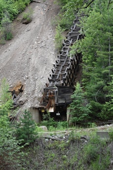 An abandoned, old wooden mining structure is situated on a steep, rocky hillside, partially surrounded by lush green foliage and trees.