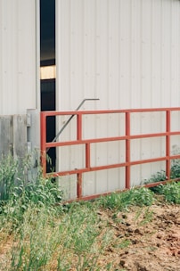 A metal gate painted red stands partially in front of a large white industrial building. The gate is open, revealing a dark interior with a faint glow of light inside. Tall grass and some foliage grow around the gate and building, adding a rustic touch to the scene.