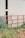 A metal gate painted red stands partially in front of a large white industrial building. The gate is open, revealing a dark interior with a faint glow of light inside. Tall grass and some foliage grow around the gate and building, adding a rustic touch to the scene.