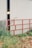 A metal gate painted red stands partially in front of a large white industrial building. The gate is open, revealing a dark interior with a faint glow of light inside. Tall grass and some foliage grow around the gate and building, adding a rustic touch to the scene.