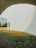 A modern architectural scene with a large white curved building and a green lawn in front. A person is seated at a table under the building's shade, surrounded by lush grass and open space. The sky is clear, contributing to the bright and serene atmosphere.