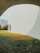 A modern architectural scene with a large white curved building and a green lawn in front. A person is seated at a table under the building's shade, surrounded by lush grass and open space. The sky is clear, contributing to the bright and serene atmosphere.