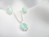 A set of jewelry featuring a necklace and matching earrings, each adorned with light blue gemstones encircled by small silver embellishments, displayed on a textured white fabric background.