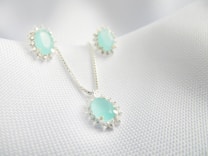 A set of jewelry featuring a necklace and matching earrings, each adorned with light blue gemstones encircled by small silver embellishments, displayed on a textured white fabric background.