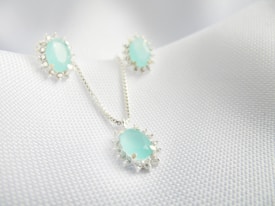 A set of jewelry featuring a necklace and matching earrings, each adorned with light blue gemstones encircled by small silver embellishments, displayed on a textured white fabric background.