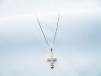 A delicate silver necklace with a cross-shaped pendant, resting on a soft white textured fabric. The pendant is studded with small sparkling stones, adding a touch of elegance.