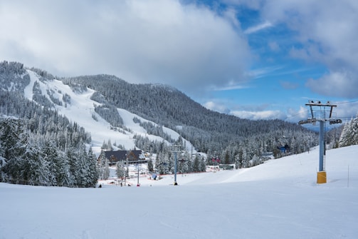 A snowy mountain landscape featuring a ski resort. The image includes ski slopes surrounded by dense forests covered in snow. A ski lift is visible on the right, extending up the mountain. A few buildings and people can be seen at the base of the slopes, likely part of the resort. The sky is mostly cloudy with patches of blue.
