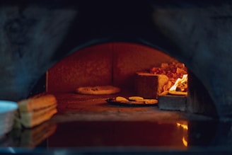 A stone pizza oven with a visible fire on the right side heating the space. Inside, there are a few pieces of bread or baked goods on a tray and a single flatbread or pizza positioned directly on the oven's stone surface. The interior is warmly lit by the orange glow of the flames.