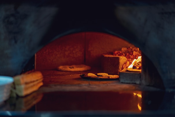 A stone pizza oven with a visible fire on the right side heating the space. Inside, there are a few pieces of bread or baked goods on a tray and a single flatbread or pizza positioned directly on the oven's stone surface. The interior is warmly lit by the orange glow of the flames.