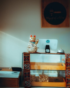 A small display counter with a glass case contains two trays of pastries. On top of the counter is a golden waving cat figurine, often seen as a symbol of luck. A tablet with a stand is used as a point-of-sale system, alongside a metal service bell. The counter also features a decorative cloth with intricate patterns.