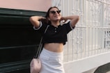 A person wearing sunglasses and casual clothing stands in front of a decorative white metal fence and dark green wall. They have their hands behind their head and are smiling, conveying a relaxed and carefree demeanor. The outfit includes a black cropped polo shirt and a white skirt, accessorized with a light pink handbag with a chain strap.