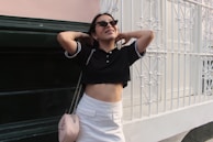 A person wearing sunglasses and casual clothing stands in front of a decorative white metal fence and dark green wall. They have their hands behind their head and are smiling, conveying a relaxed and carefree demeanor. The outfit includes a black cropped polo shirt and a white skirt, accessorized with a light pink handbag with a chain strap.