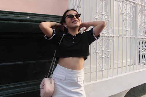 A person wearing sunglasses and casual clothing stands in front of a decorative white metal fence and dark green wall. They have their hands behind their head and are smiling, conveying a relaxed and carefree demeanor. The outfit includes a black cropped polo shirt and a white skirt, accessorized with a light pink handbag with a chain strap.