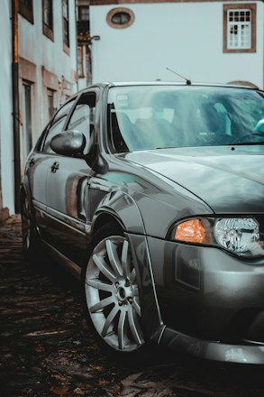 A sleek, silver car is parked on a cobblestone street in a residential area. The setting has a somewhat vintage look, with older buildings visible in the background. The car is reflected clearly on its shiny surface, indicating it is well-maintained. The composition highlights the front wheel and headlight, suggesting a focus on the car&rsquo;s design.