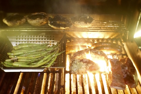 A barbecue grill with a variety of food items being cooked. There are asparagus spears in a perforated metal tray, sausages on the grill, and pieces of meat that are sizzling under a bright flame. Portobello mushrooms are grilled on a rack above the main surface.