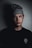 A young person wearing a knitted gray beanie and a black t-shirt with a logo featuring a sun design. The individual has facial and neck tattoos, including a small skull on the neck and numerical designs on the face. The background is dimly lit, casting shadows on the right side of the face.