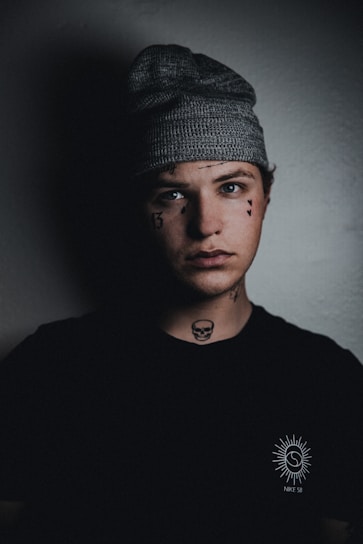A young person wearing a knitted gray beanie and a black t-shirt with a logo featuring a sun design. The individual has facial and neck tattoos, including a small skull on the neck and numerical designs on the face. The background is dimly lit, casting shadows on the right side of the face.