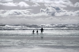 A family of three walks along the shore of a beach, with waves crashing in the background. The sky is filled with fluffy clouds, and the water reflects the light, creating a shimmering effect.