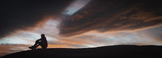 a man sitting on top of a hill under a cloudy sky