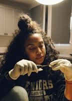 A young woman with curly hair tied in a bun is wearing gloves while carefully handling a tool in a softly lit room. Her attire has text indicating 'Seniors Class Of' and she appears focused and absorbed in her task.