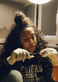 A young woman with curly hair tied in a bun is wearing gloves while carefully handling a tool in a softly lit room. Her attire has text indicating 'Seniors Class Of' and she appears focused and absorbed in her task.