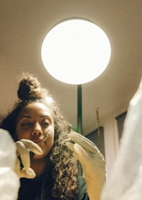 A person with curly hair and a focused expression is wearing gloves and using a small tool under a bright, circular light. The scene suggests a meticulous task, possibly related to science or art.