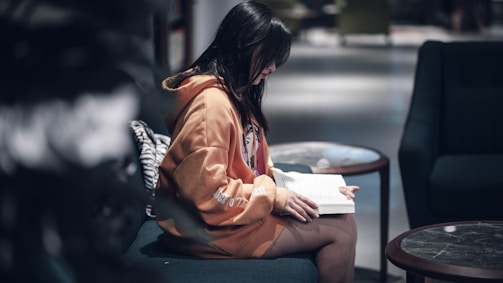 A person with long dark hair is sitting on a blue sofa, wearing an orange hoodie, and reading a book. The setting appears to be a cozy indoor environment with soft lighting, and there are round wooden tables nearby.