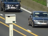 Two cars are seen traveling on a highway, one is a Nissan and the other is a BMW. A traffic camera is positioned beside the road, monitoring passing vehicles. The highway has clear yellow lines and a concrete barrier separates the lanes.