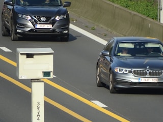 Two cars are seen traveling on a highway, one is a Nissan and the other is a BMW. A traffic camera is positioned beside the road, monitoring passing vehicles. The highway has clear yellow lines and a concrete barrier separates the lanes.