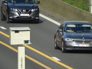 Two cars are seen traveling on a highway, one is a Nissan and the other is a BMW. A traffic camera is positioned beside the road, monitoring passing vehicles. The highway has clear yellow lines and a concrete barrier separates the lanes.