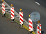 A row of traffic barriers with red and white diagonal stripes is arranged along the edge of a road. Each barrier is topped with an orange warning light. Beside the barriers, there is a blank circular sign mounted on a pole. The road surface is dark with a section marked by a solid white line, and there are scattered leaves on the ground.