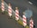 A row of traffic barriers with red and white diagonal stripes is arranged along the edge of a road. Each barrier is topped with an orange warning light. Beside the barriers, there is a blank circular sign mounted on a pole. The road surface is dark with a section marked by a solid white line, and there are scattered leaves on the ground.