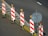 A row of traffic barriers with red and white diagonal stripes is arranged along the edge of a road. Each barrier is topped with an orange warning light. Beside the barriers, there is a blank circular sign mounted on a pole. The road surface is dark with a section marked by a solid white line, and there are scattered leaves on the ground.