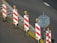 A row of traffic barriers with red and white diagonal stripes is arranged along the edge of a road. Each barrier is topped with an orange warning light. Beside the barriers, there is a blank circular sign mounted on a pole. The road surface is dark with a section marked by a solid white line, and there are scattered leaves on the ground.
