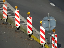 A row of traffic barriers with red and white diagonal stripes is arranged along the edge of a road. Each barrier is topped with an orange warning light. Beside the barriers, there is a blank circular sign mounted on a pole. The road surface is dark with a section marked by a solid white line, and there are scattered leaves on the ground.