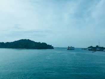 A serene seascape with a body of water in the foreground and a small island covered in green vegetation in the distance. A ferry is sailing to the right, approaching a jetty where several buildings and towers are visible. The sky is light with scattered clouds, creating a calm atmosphere.