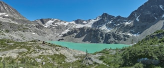 A breathtaking alpine landscape featuring turquoise lake bordered by steep, rocky mountains with patches of snow. Green vegetation covers the lower slopes, while the sky is clear and blue.