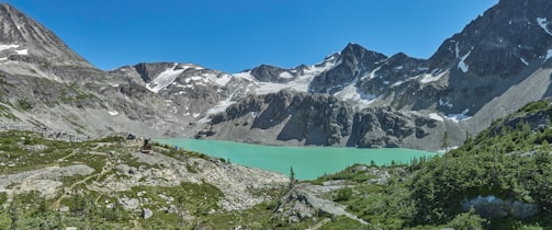 A breathtaking alpine landscape featuring turquoise lake bordered by steep, rocky mountains with patches of snow. Green vegetation covers the lower slopes, while the sky is clear and blue.
