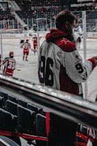 A hockey game is taking place with players in red and white uniforms skating on the ice. One player with the number 96 on their jersey is standing near the rink, possibly a coach or team member. The rink is surrounded by empty seats, and the atmosphere seems focused and dynamic.