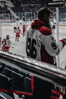 A hockey game is taking place with players in red and white uniforms skating on the ice. One player with the number 96 on their jersey is standing near the rink, possibly a coach or team member. The rink is surrounded by empty seats, and the atmosphere seems focused and dynamic.