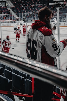 A hockey game is taking place with players in red and white uniforms skating on the ice. One player with the number 96 on their jersey is standing near the rink, possibly a coach or team member. The rink is surrounded by empty seats, and the atmosphere seems focused and dynamic.