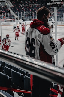 A hockey game is taking place with players in red and white uniforms skating on the ice. One player with the number 96 on their jersey is standing near the rink, possibly a coach or team member. The rink is surrounded by empty seats, and the atmosphere seems focused and dynamic.