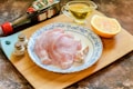 A wooden cutting board holds a plate with two raw chicken breast fillets and garlic cloves. Nearby, there is a halved orange, a small container of oil, a bottle of soy sauce, and two corked spice jars.