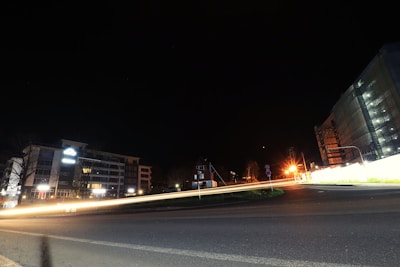 A nighttime urban scene featuring buildings on either side of a road. The road displays light streaks from passing vehicles, indicating a long exposure photograph. The sky is dark, emphasizing the artificial lights illuminating the area from streetlights and building lights.
