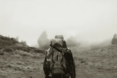 grayscale photo of man in backpack walking on dirt road