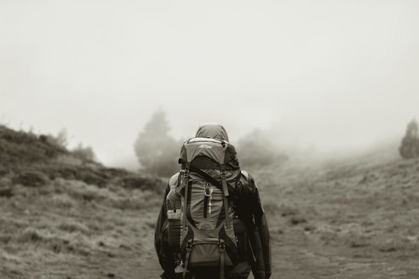 A solitary hiker with a large backpack walks along a foggy trail surrounded by mist-covered hills. The atmosphere is serene and quiet, with trees barely visible through the fog.