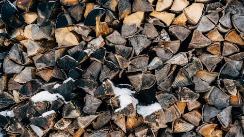 A neatly stacked pile of chopped firewood, with a dusting of snow scattered on top of the logs. The wood pieces vary in shade from light to dark brown, and the shapes are irregular, creating a textured and rustic appearance.