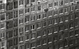 A large residential building with numerous rectangular windows, each window having a uniform appearance. The facade is weathered and monochromatic, creating a sense of repetition and uniformity. A few windows have curtains or items visible, and some air conditioning units are attached to the walls.
