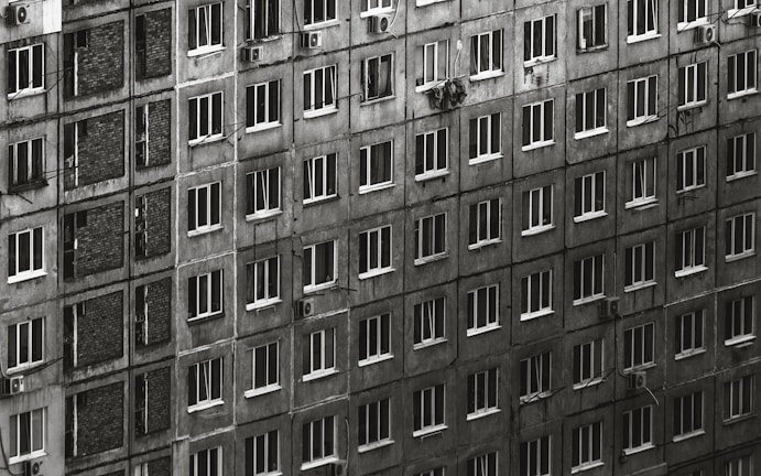 A large residential building with numerous rectangular windows, each window having a uniform appearance. The facade is weathered and monochromatic, creating a sense of repetition and uniformity. A few windows have curtains or items visible, and some air conditioning units are attached to the walls.