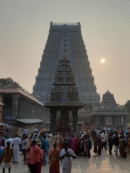 A large, intricately carved Hindu temple tower dominates the scene against the backdrop of a hazy sky with a visible sun. The foreground is bustling with people, many of whom appear to be worshippers or tourists, walking around and interacting near the temple.